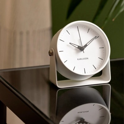 Et stilfuldt analogt vækkeur fra Karlsson med hvid urskive og sorte visere. Uret har et minimalistisk design med tynde timemarkeringer og en skrå metalramme i hvid, som giver det et moderne look. Det står på en glasbordplade og spejler sig i overfladen, mens en grøn plante og gardiner er synlige i baggrunden.