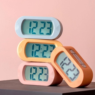 yra färgglada digitala väckarklockor staplade på varandra mot en rosa bakgrund. Klockorna har en oval design med stora LCD-skärmar som visar tiden i blå siffror. De finns i pastellfärger såsom ljusblå, gul, rosa och orange