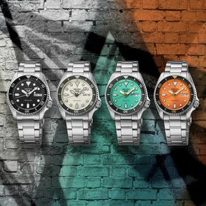 yra Seiko 5 Sports Midi klockor med stållänkar och färgade urtavlor i svart, beige, turkos och orange, mot en bakgrund av en graffitimålning i tegel.