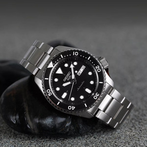 Seiko 5 Sports dykkerinspirert klokke med svart urskive, hvite selvlysende markeringer og en dagsdatovisning klokken 3. Klokken har en robust stålkonstruksjon, en roterende lunette og et klassisk stålarmbånd som kombinerer funksjonalitet og stil. Innfattet i glatte steiner med en strukturert bakgrunn som understreker den sporty og slitesterke designen.