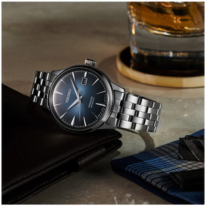 Seiko Presage herreklokke med en dypblå urskive med solinnstråling, sølvfargede visere og markører og datovisning ved klokken 3. Klokken har en elegant stålkasse og et klassisk femradet stålarmbånd som understreker det sofistikerte designet. Sett mot en stilfull bakgrunn med en skinnlommebok, mansjettknapper og et glass whisky, som understreker urets tidløse eleganse og raffinerte stil.
