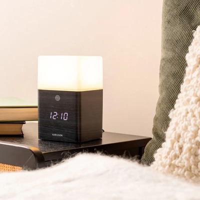 Et moderne vækkeur med indbygget lampe fra Karlsson, placeret på et natbord. Uret har en mørk trælignende base med en digital tidsvisning i hvid LED og en øvre del, der fungerer som en blød lyskilde. En bog og en grøn pude er synlige i baggrunden, mens et hvidt tæppe er i forgrunden og skaber et hyggeligt soveværelsesmiljø.